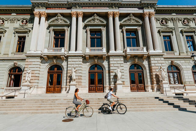 Low angle shot of a man and a woman cycling in front of the Grand Théâtre de Genève